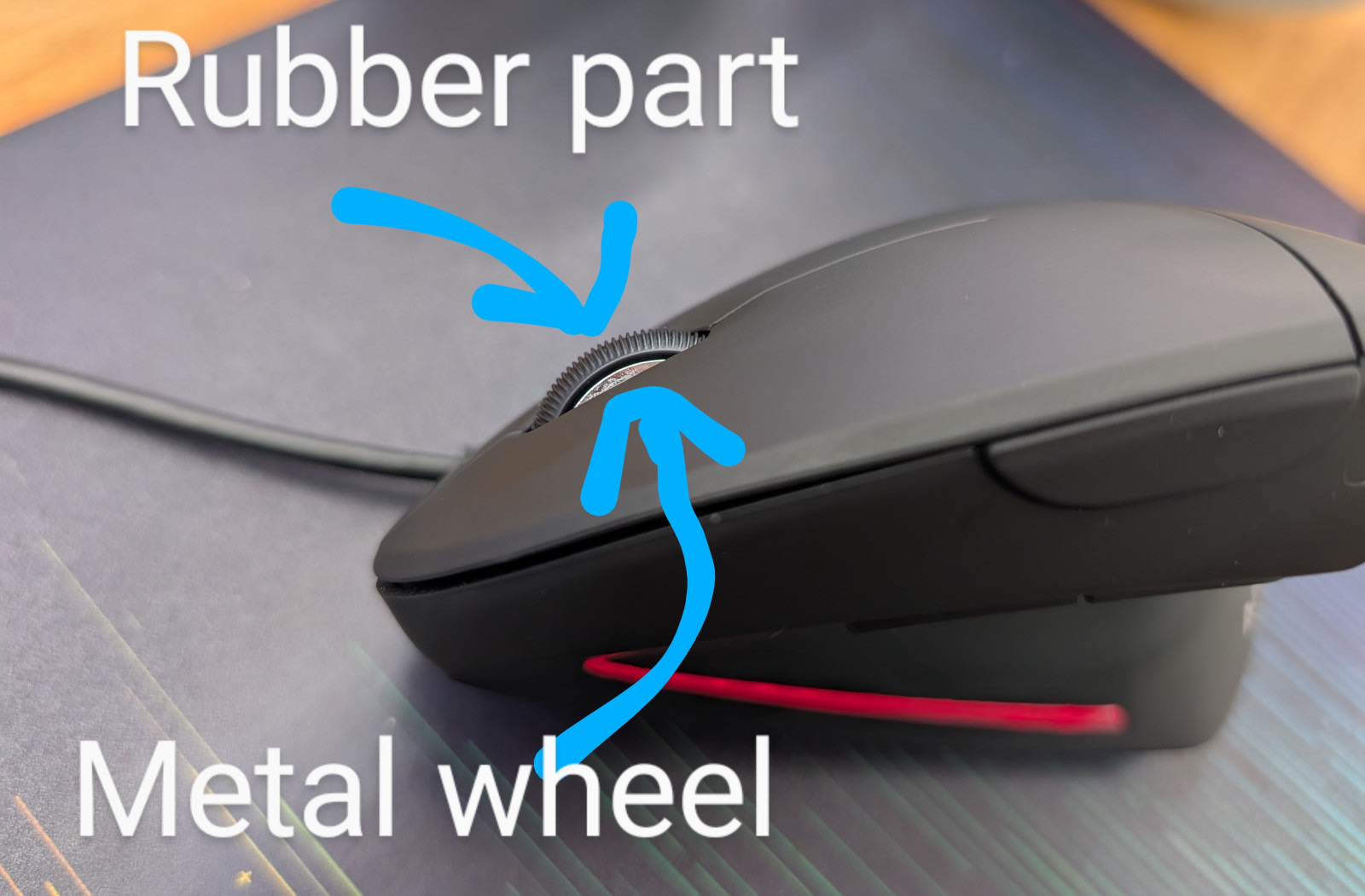 Close-up of a black Anker vertical mouse on a desk with blue annotation arrows and white text labels. The top arrow points to the black textured rubber ring around the scroll wheel labeled "Rubber part". The bottom arrow points to the silver ridged metal wheel visible beneath, labeled "Metal wheel".